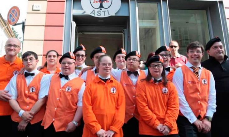 Quinze pessoas com Síndrome de Down, dentre homens e mulheres, de uniforme laranja do hotel onde trabalham, posando em frente à fachada do hotel. Apenas um deles usa uma roupa preta