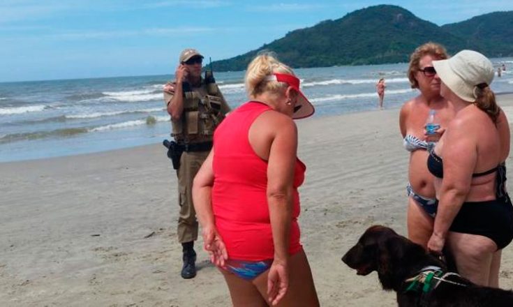 Foto em uma praia, com um policial de frente e três senhoras, de costas, e um cão, todos na areia. Ao fundo, o mar.
