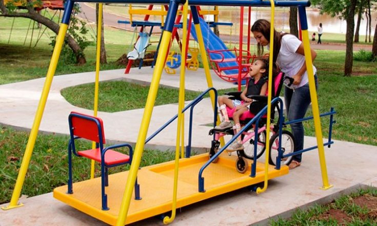 Parque de diversão em que uma garotinha, em uma cadeira de rodas, brinca em um balanço amarelo adaptado, empurrada por uma mulher