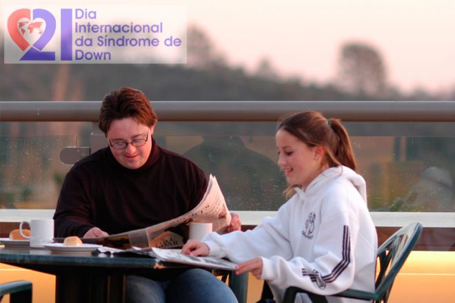 Um casal, ele com Síndrome de Down, sentado em uma mesa, tomando café, lendo jornal e com montanhas ao fundo. No alto, à esquerda, a logomarca do Dia Internacional da Síndrome de Down.