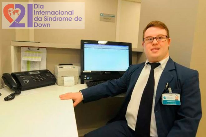 Um jovem de paletó e gravata, com Síndrome de Down, em um escritório, com um computador e um aparelho de fax a sua frente. No alto, à esquerda, a logomarca do Dia Internacional da Síndrome de Down.