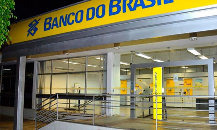Visão frontal de uma agência do Banco do Brasil, vazia, com a placa amarela e a logomarca e o nome Banco do Brasil, em azul, na parte superior.