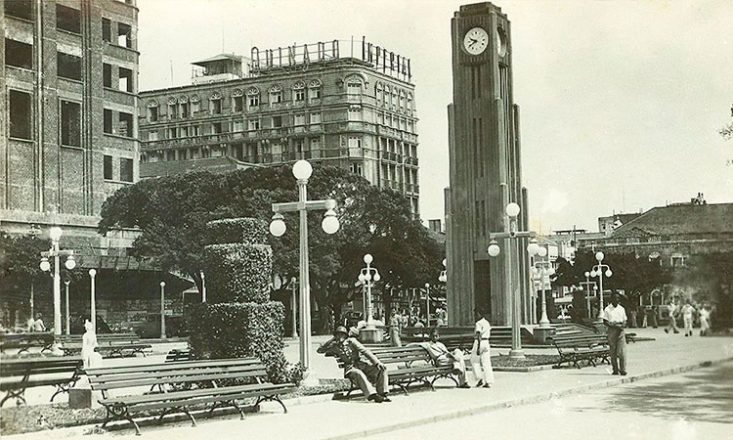 Foto em preto e branco da Praça do Ferreira, com a torre do relógio à direita, prédios ao fundo, vários bancos e pessoas caminhando ou sentadas