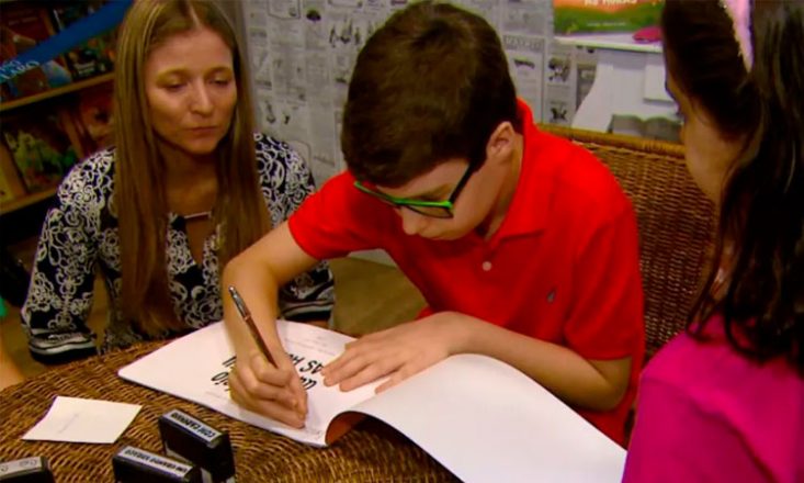 Um garoto autista de 10 anos, ao centro, assina um livro escrito por ele. Ao seu lado, sua mãe o observa e orienta. Do outro lado, uma garotinha o aguarda