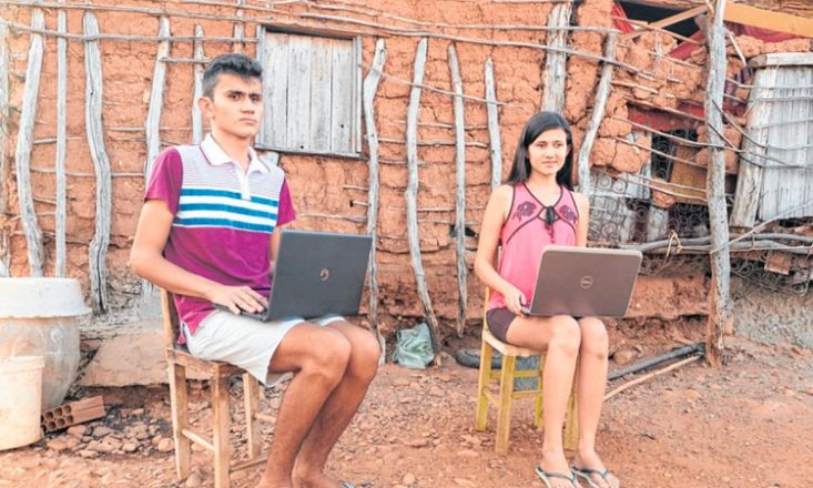 Dois irmãos, um rapaz (à esquerda) e uma moça, sentados em cadeiras de madeira, com um laptop no colo, no terreiro de uma casa bastante humilde