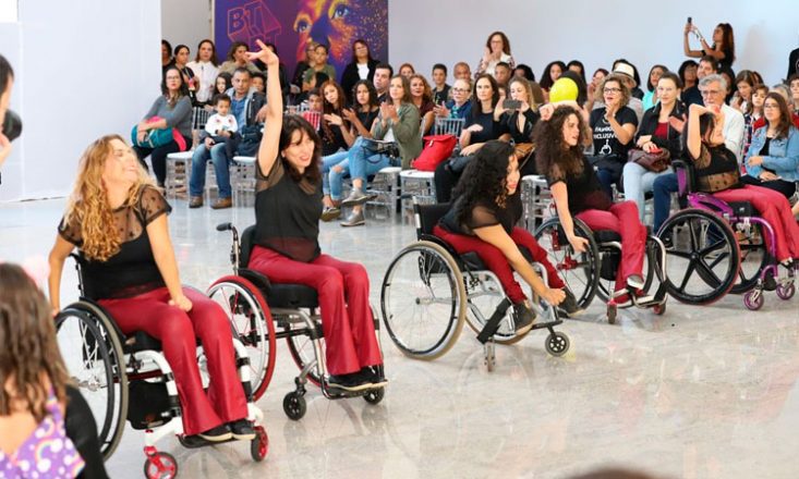 Cinco moças cadeirantes dançam em uma apresentação cultural para uma platéia composta de homens e mulheres ao seu redor.