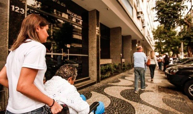 Foto horizontal de uma calçada do Rio de Janeiro, com uma jovem empurrando a cadeira de rodas de um idoso, em primeiro plano, e algumas pessoas caminhando mais a frente.