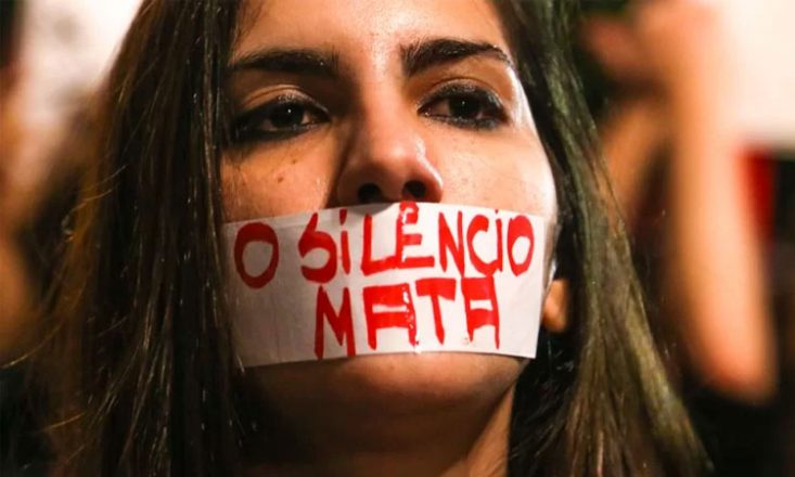 Rosto de uma mulher com um adesivo na boca escrito, em letras vermelhas: O SILÊNCIO MATA.