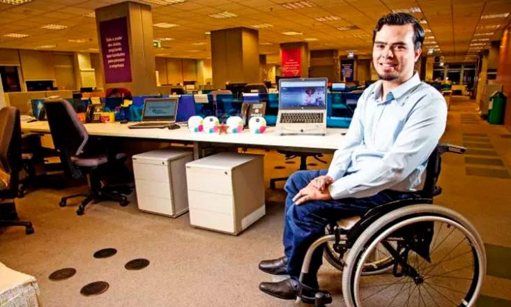 Homem em uma cadeira de rodas, de lado, sorrindo, no canto direito da imagem. À esquerda, uma mesa com dois computadores e vários enfeites.