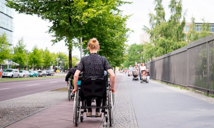 Mulher em cadeira de rodas, de costas, segue por uma rua. A sua frente, outro cadeirante. Ao fundo, algumas pessoas caminham na direção oposta. Do lado esquerdo da imagem, vários carros estacionados.